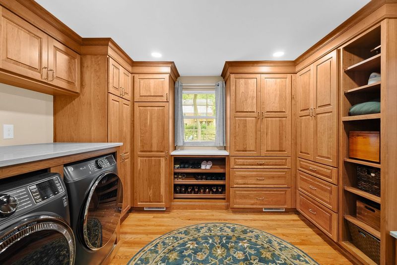 Bethel Park Custom Laundry Room with Walk-In Closet Boasts Wood Cabinetry in a Warm Natural Finish, Open Shelving for Folding Items, Baskets and Decorated Touches Blending Practicality of a Laundry Room with the Elegance of a Stylish Dressing Space.