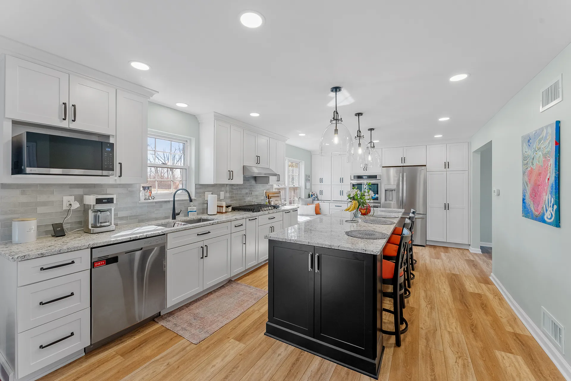 Transitional O’Hara Township Kitchen Featuring White Shaker -Style Cabinets, Warm Light and Medium Gray Countertops, Black Island and Light Wood Floor.