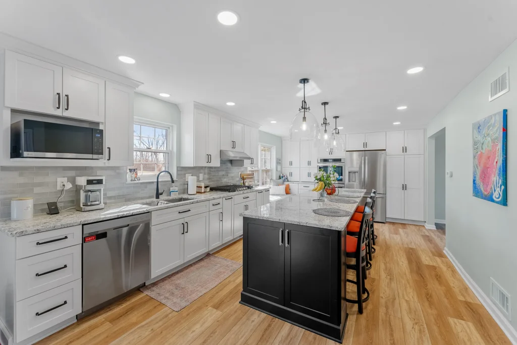 Transitional O ’ Hara T ownship Kitchen Featuring White Shaker - Style Cabinets, Warm Light and Medium Gray Countertops, Black Island and Light Wood Floor.