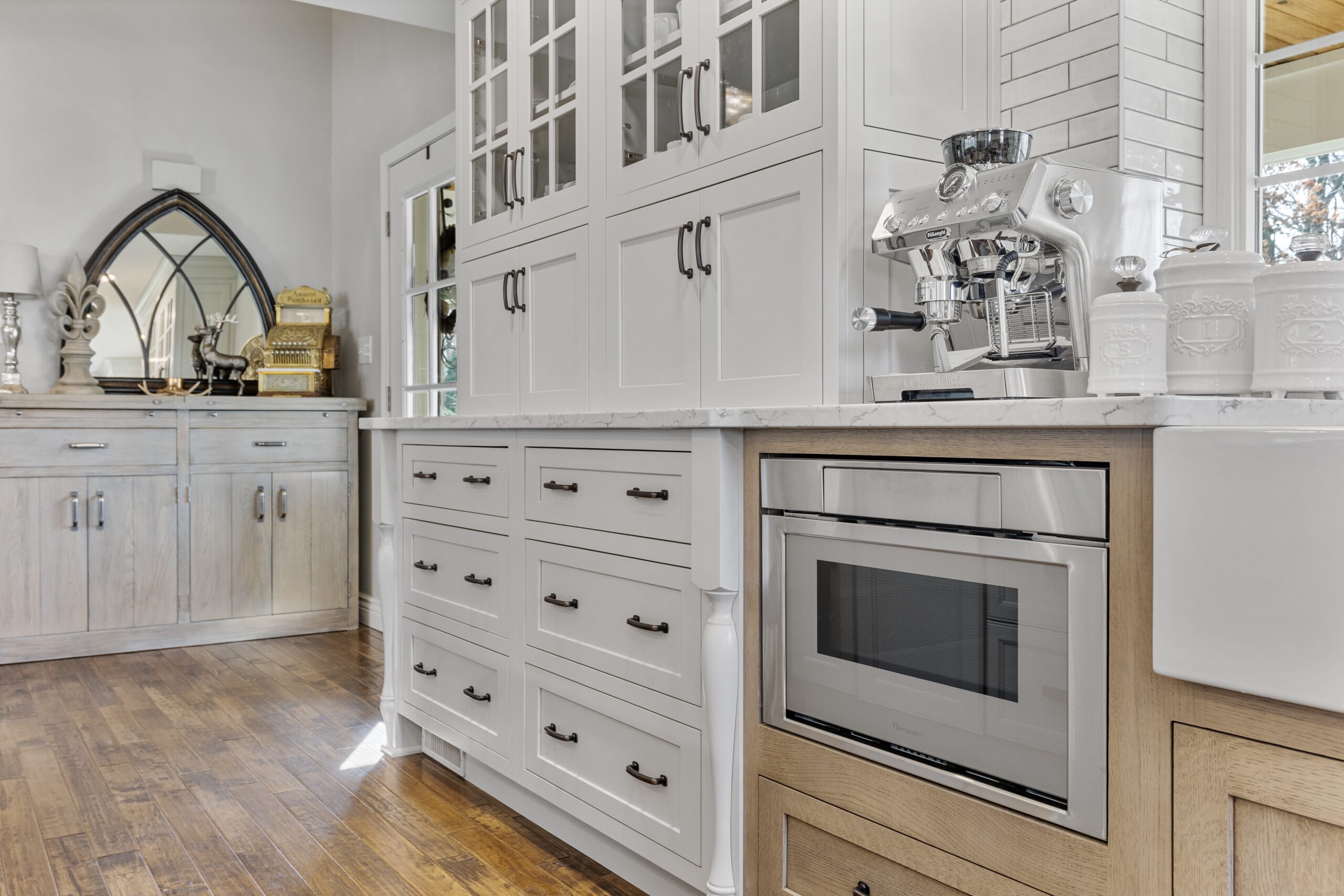 Bright Transitional Farmhouse Kitchen With Mix of White, Light Gray and Stain Shaker Cabinets, Quartz Countertop With Veining, Hardwood Floors and a Large Island.